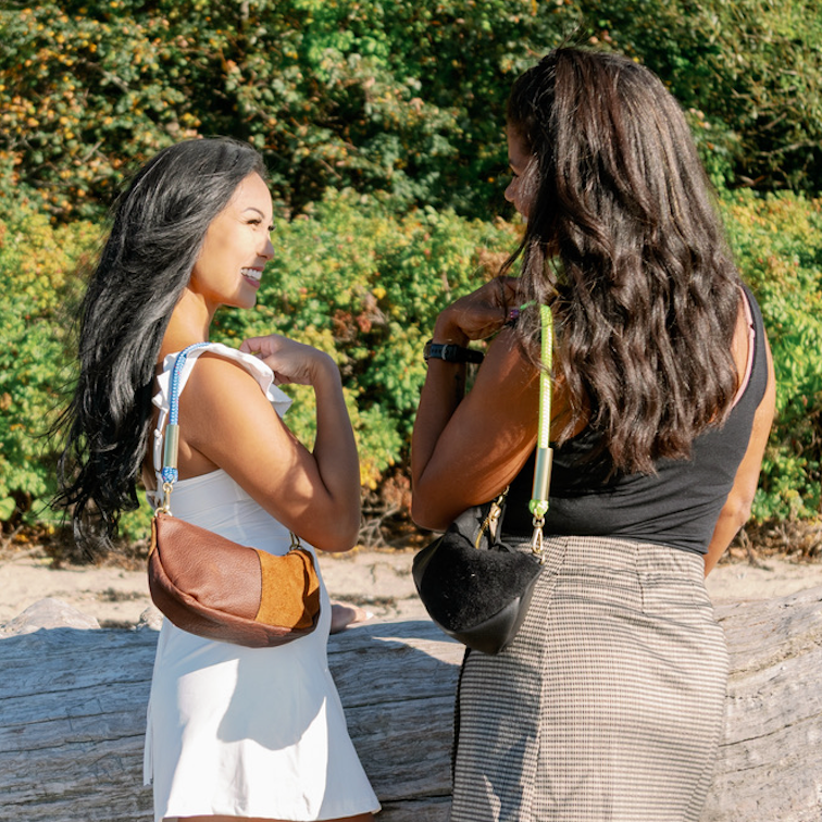 Two women standing on a sandy beach in front of a large piece of driftwood, with dense green foliage in the background. One woman wears a white sleeveless top and white skirt, and the other wears a black sleeveless top and gray skirt. They are both wearing Classic Zip bags, left has a Brown bag and the right has a Black Bag on.