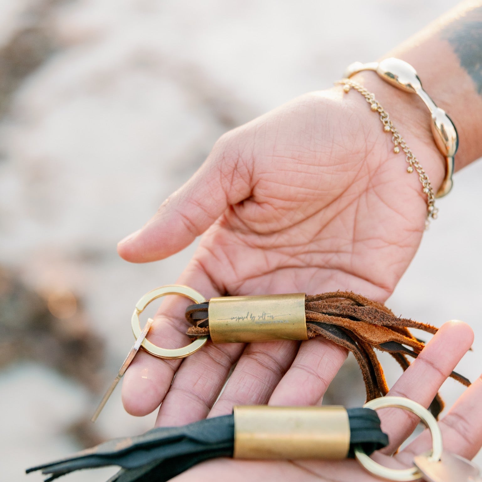 Hand holding two keychains with leather tassels against a blurred natural background