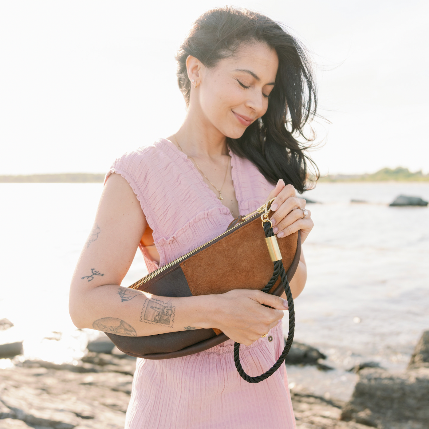 A woman in a pink ribbed dress stands on a rocky shoreline holding a brown and black clutch bag with a gold-tone zipper and black braided strap.