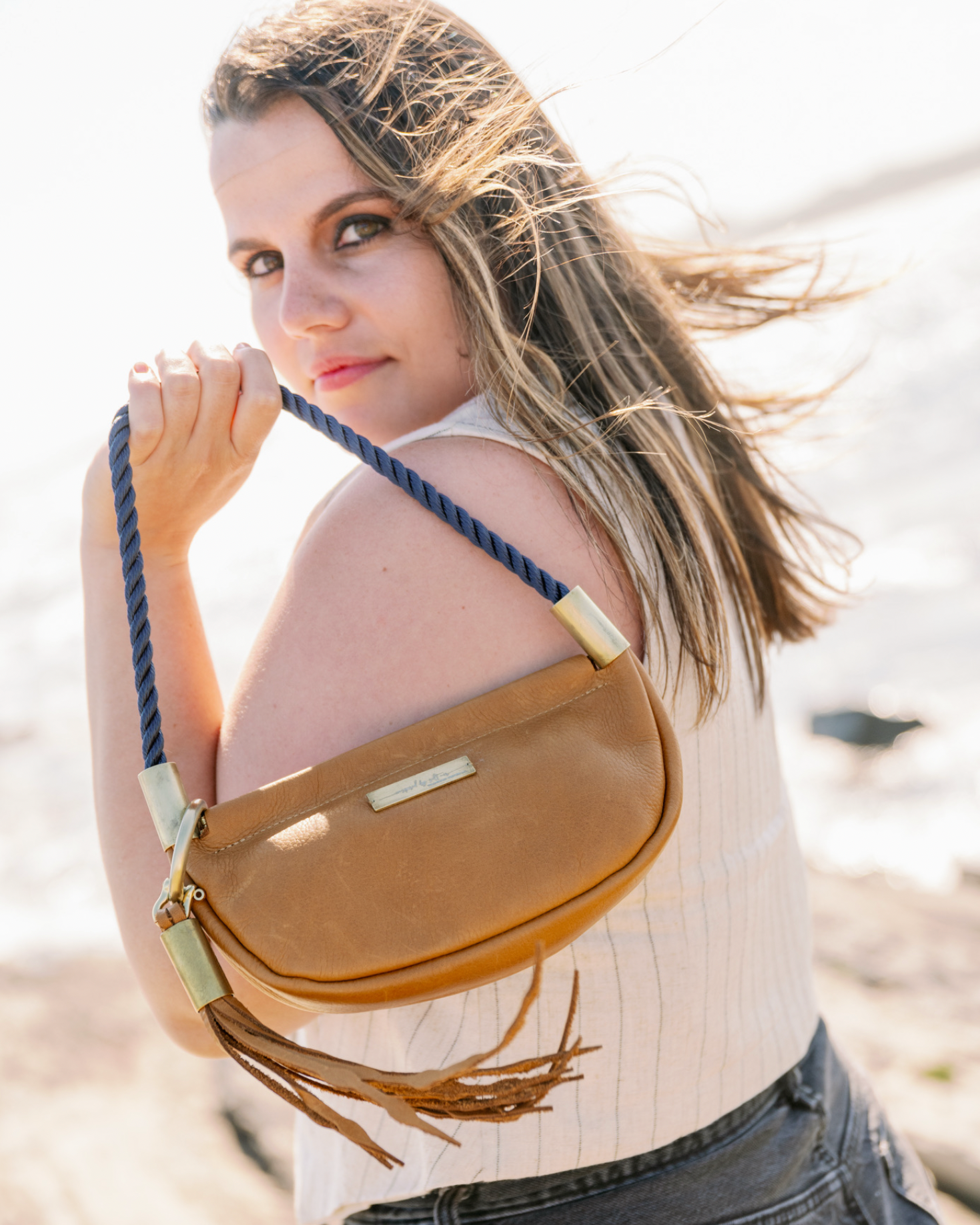 A woman on a beach wearing a tan leather crescent-shaped shoulder bag with a navy blue braided strap and gold-tone hardware.