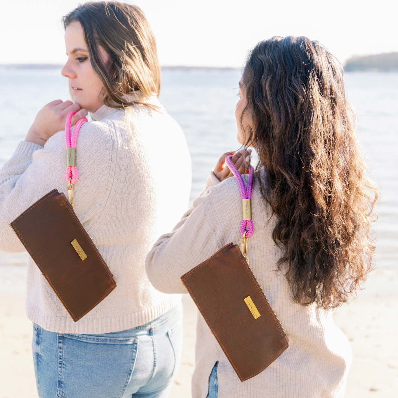 Two women standing on a beach, both wearing brown leather clutches with bright pink woven straps and gold-tone metal accents.