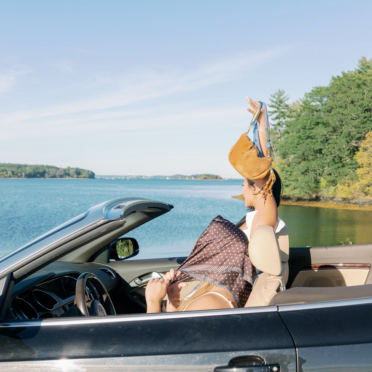 2 woman sits in a black convertible car by the water. The passenger has her arm raised in the air holding the Beach Nut Classic Zip Bag with the Harborside Blue rope. The driver is tying a scarf on her head. Trees line the shore under a clear blue sky.