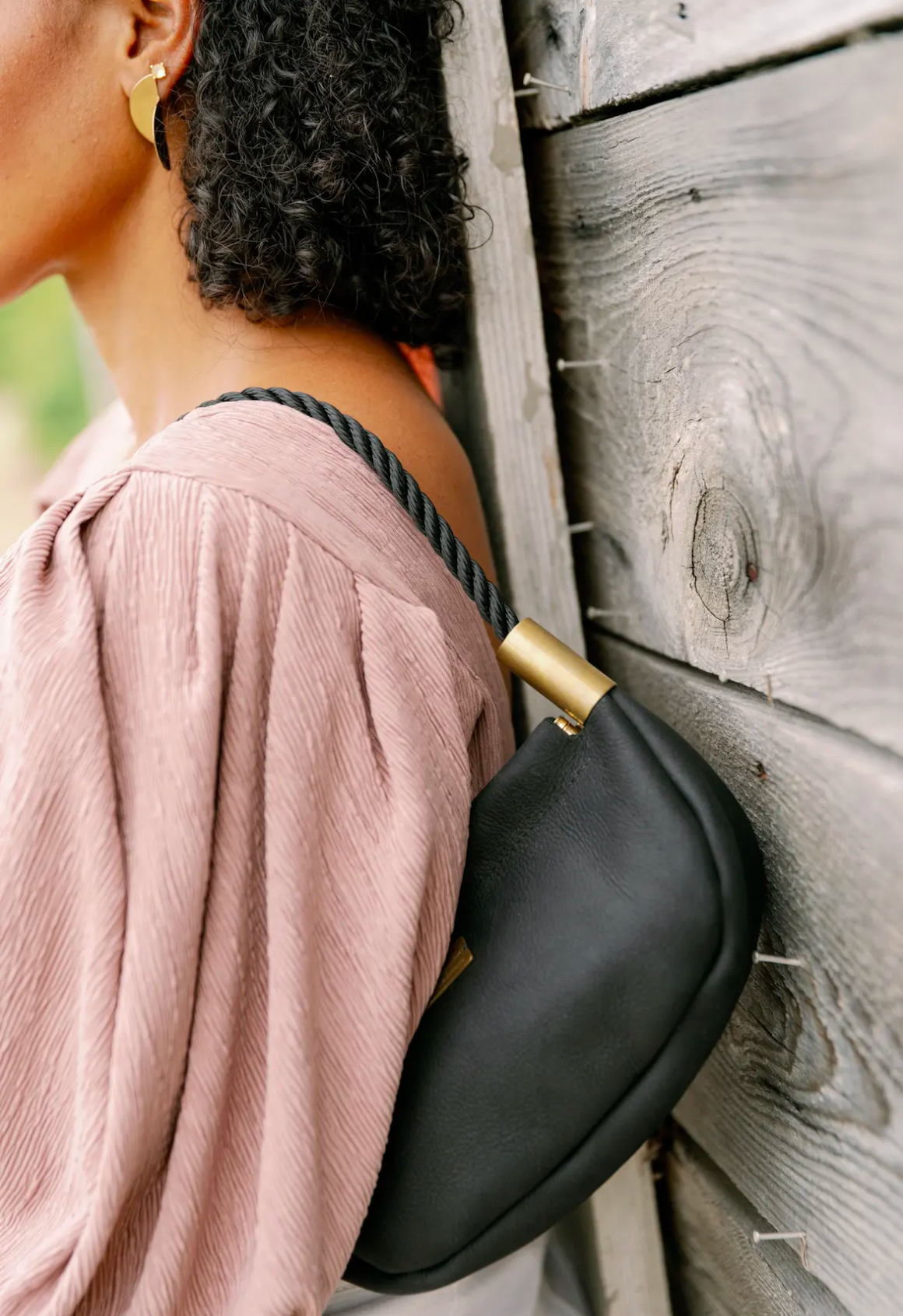 Person wearing a black crossbody bag with gold clasp against a wooden wall.