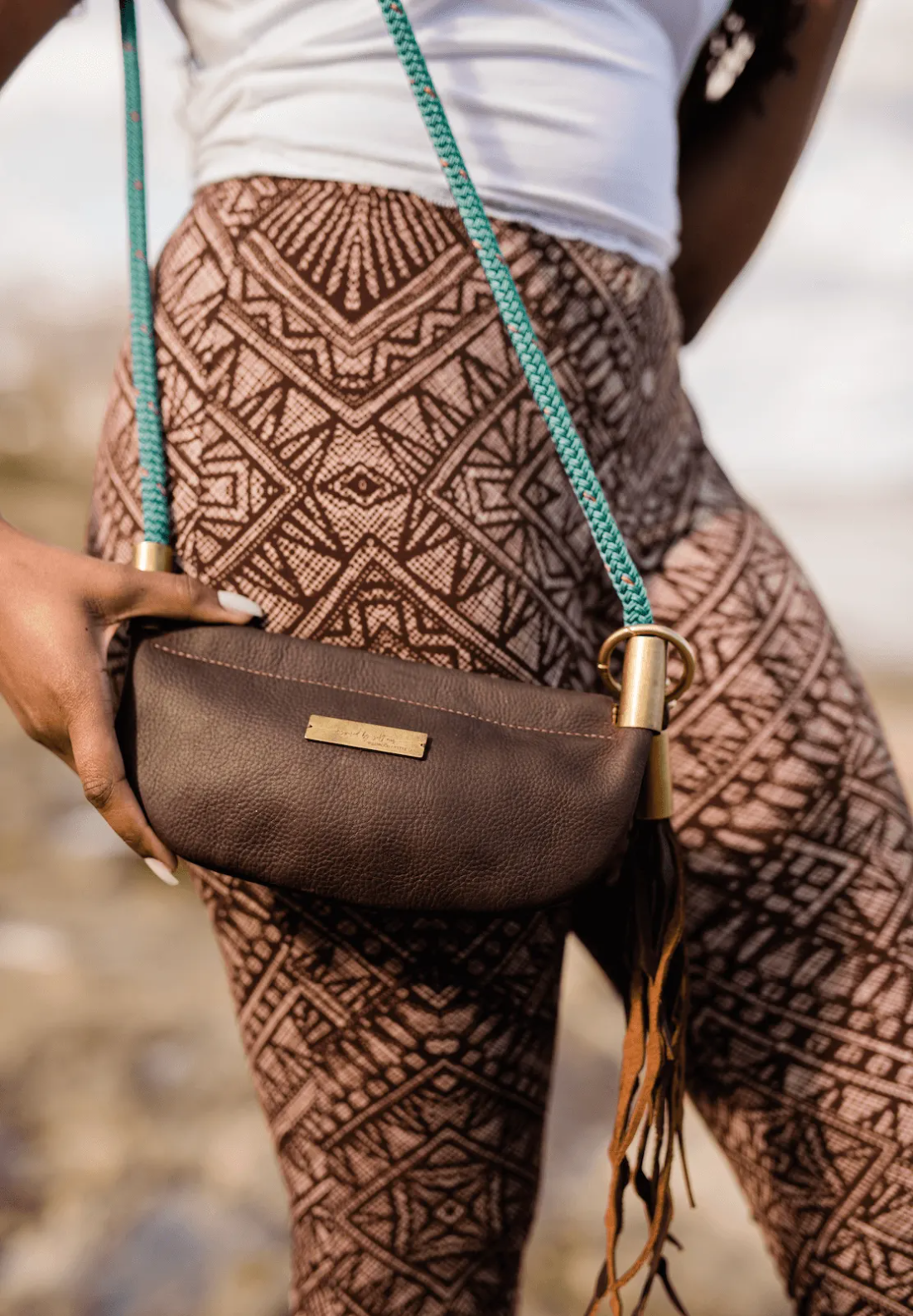 Brown leather bag with gold buckle held by a person wearing patterned pants.