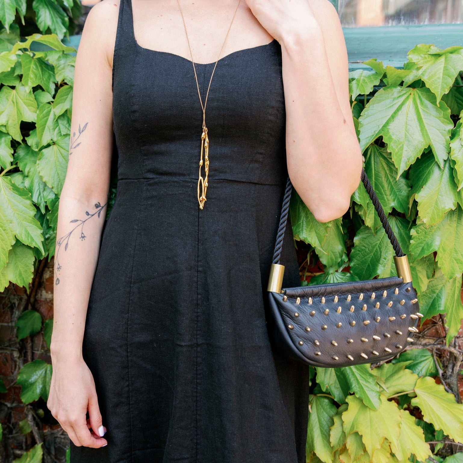Person wearing a black dress and carrying a black handbag with green foliage in the background