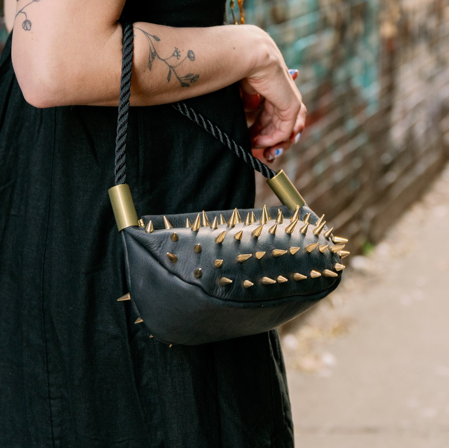 Person holding a black handbag with gold spikes against a graffiti-covered wall.