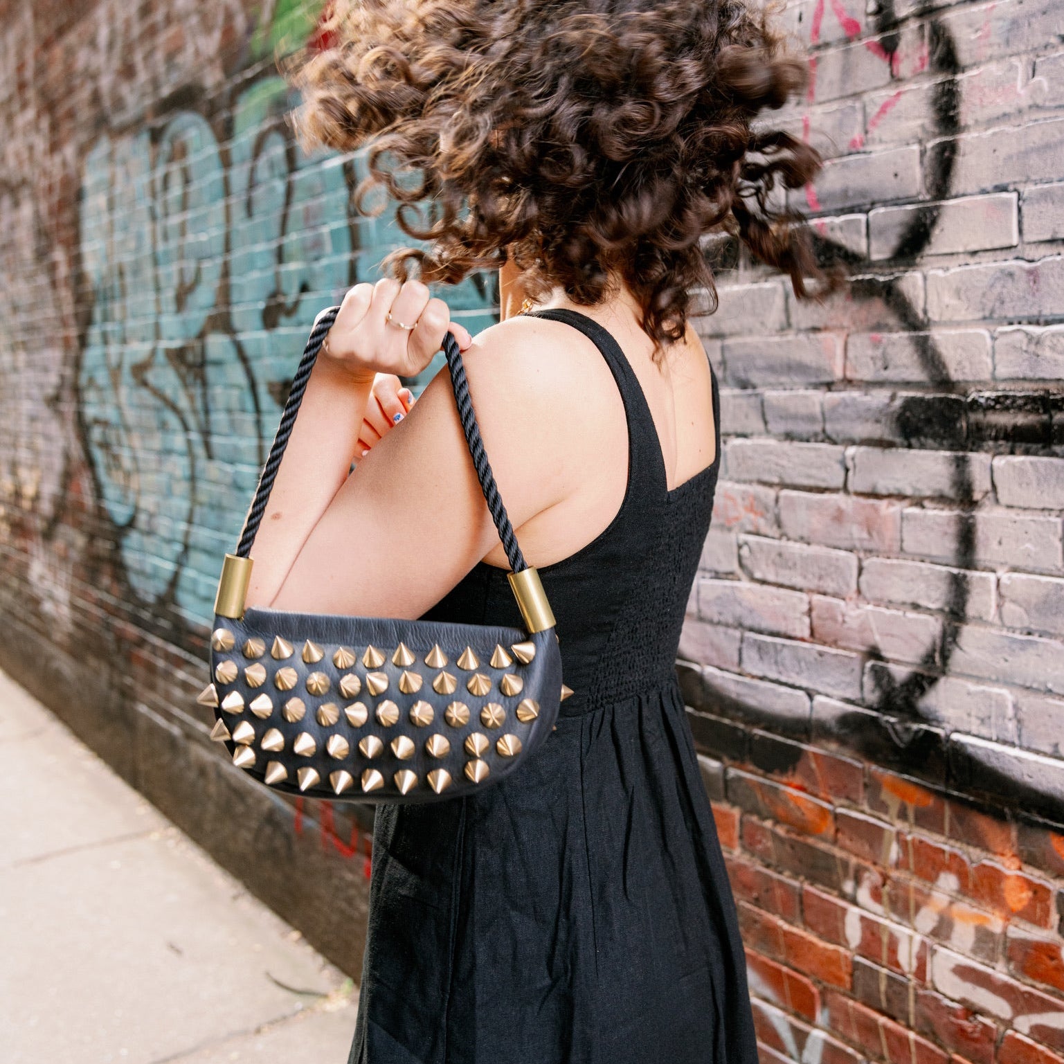 Woman in a black dress holding a studded handbag against a graffiti-covered wall.