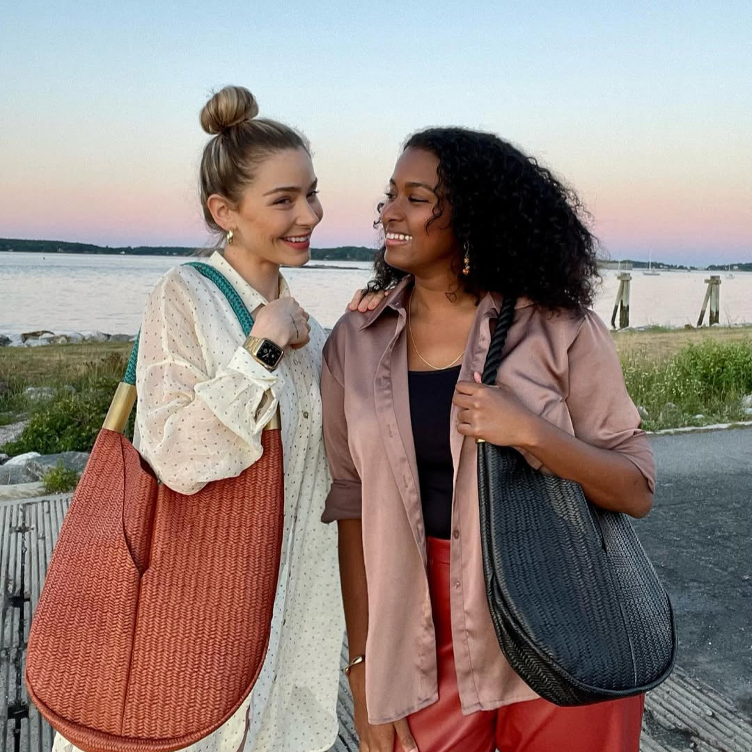 Two women standing by a waterfront, each holding a handbag, with a scenic background.