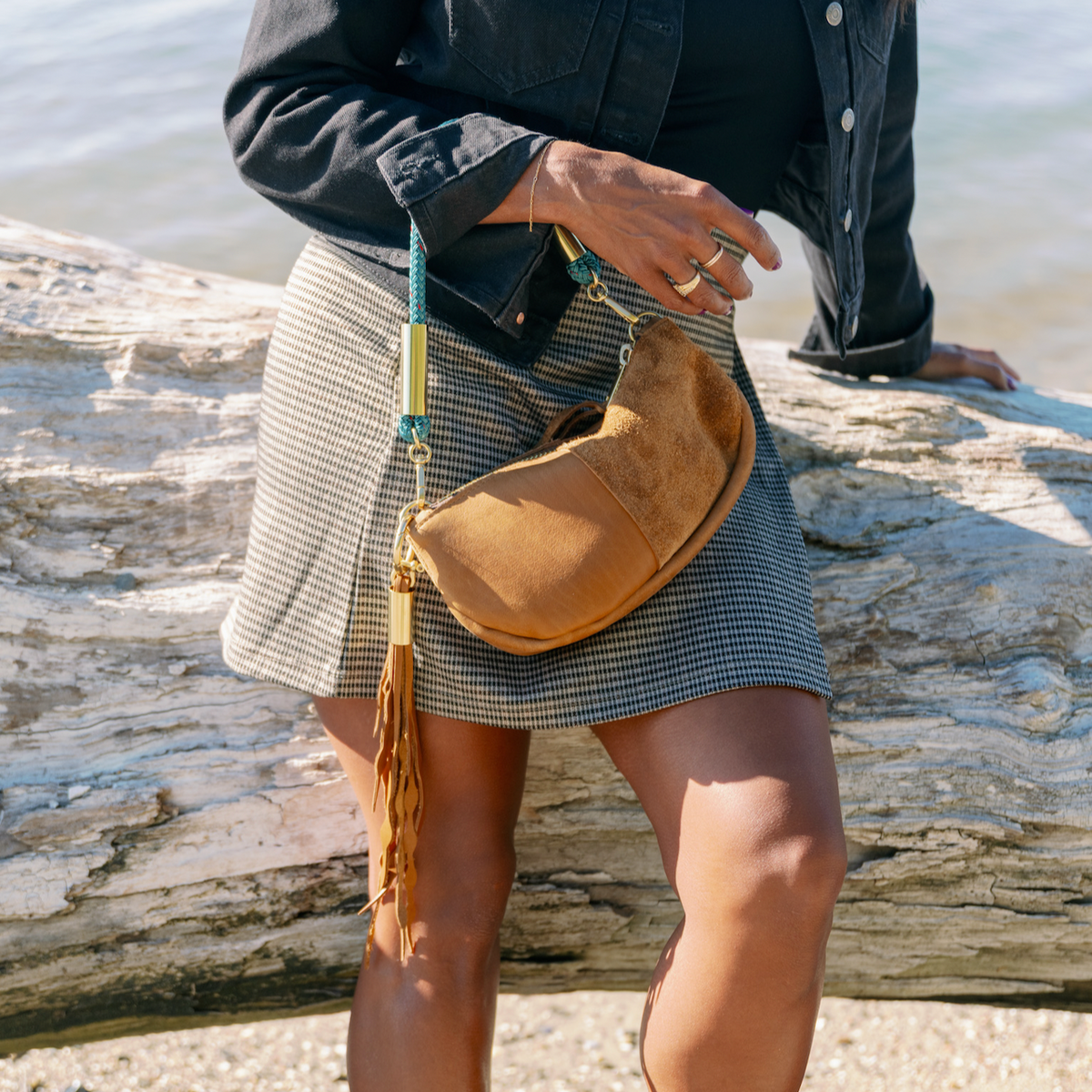 A woman at the beach leaning on a wood log, wearing the Beach Nut Convertible Zip Bag with a matching Seaweed Tassel and the Seaside Teal rope in wrist length.