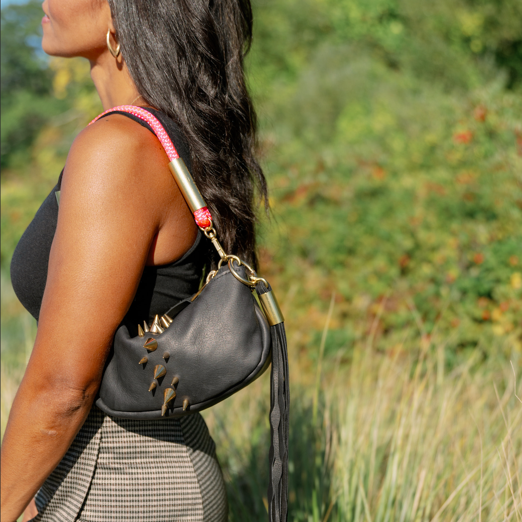 A woman in a black top and gray patterned skirt wearing a Black Sea Urchin Bag with the Neon Pink rope on her shoulder, and a Dramatic Black Seaweed Tassel, photographed outdoors in tall grass.