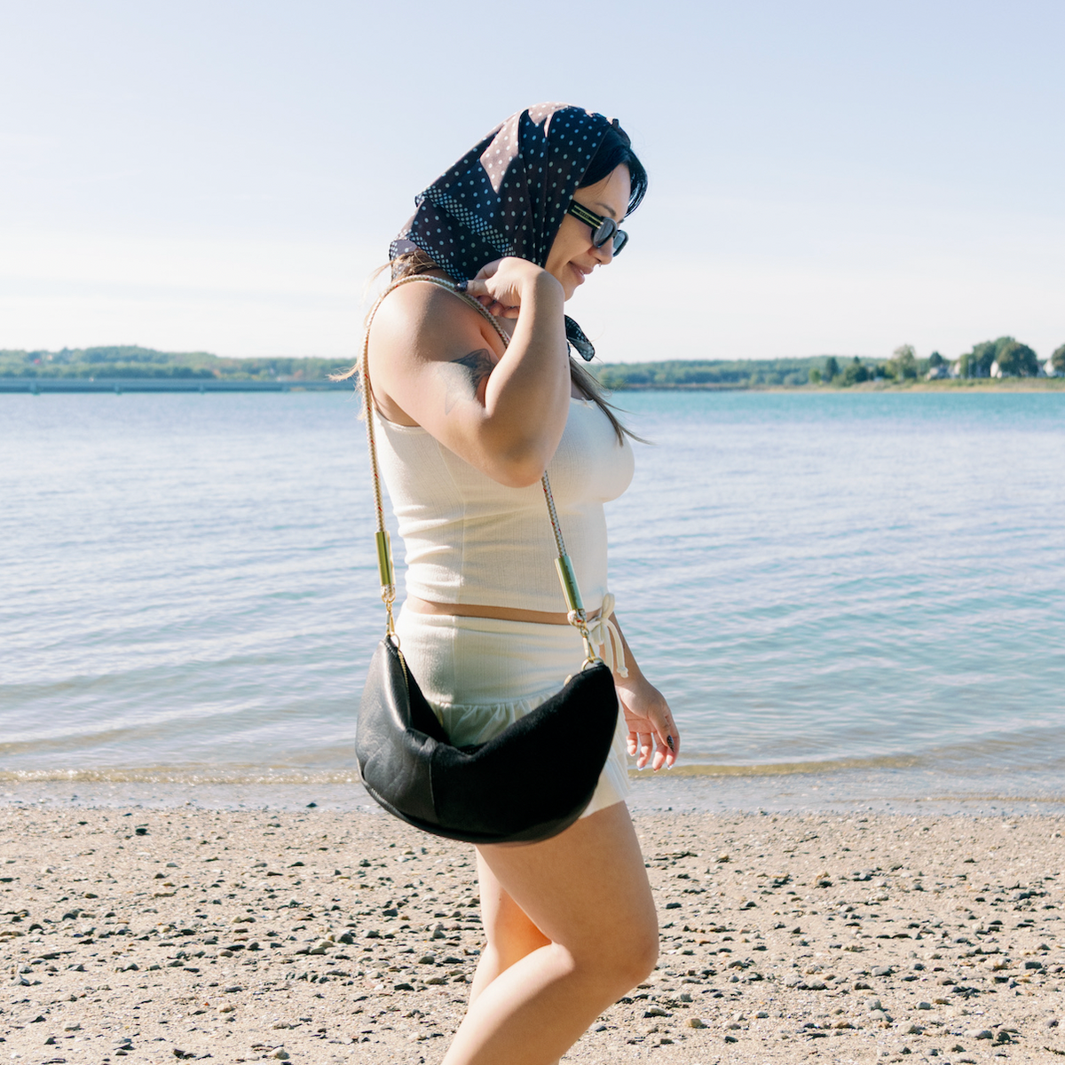 A woman walks along a pebble beach next to a lake, wearing a light yellow skirt set, and carrying a Black Sling Bag.