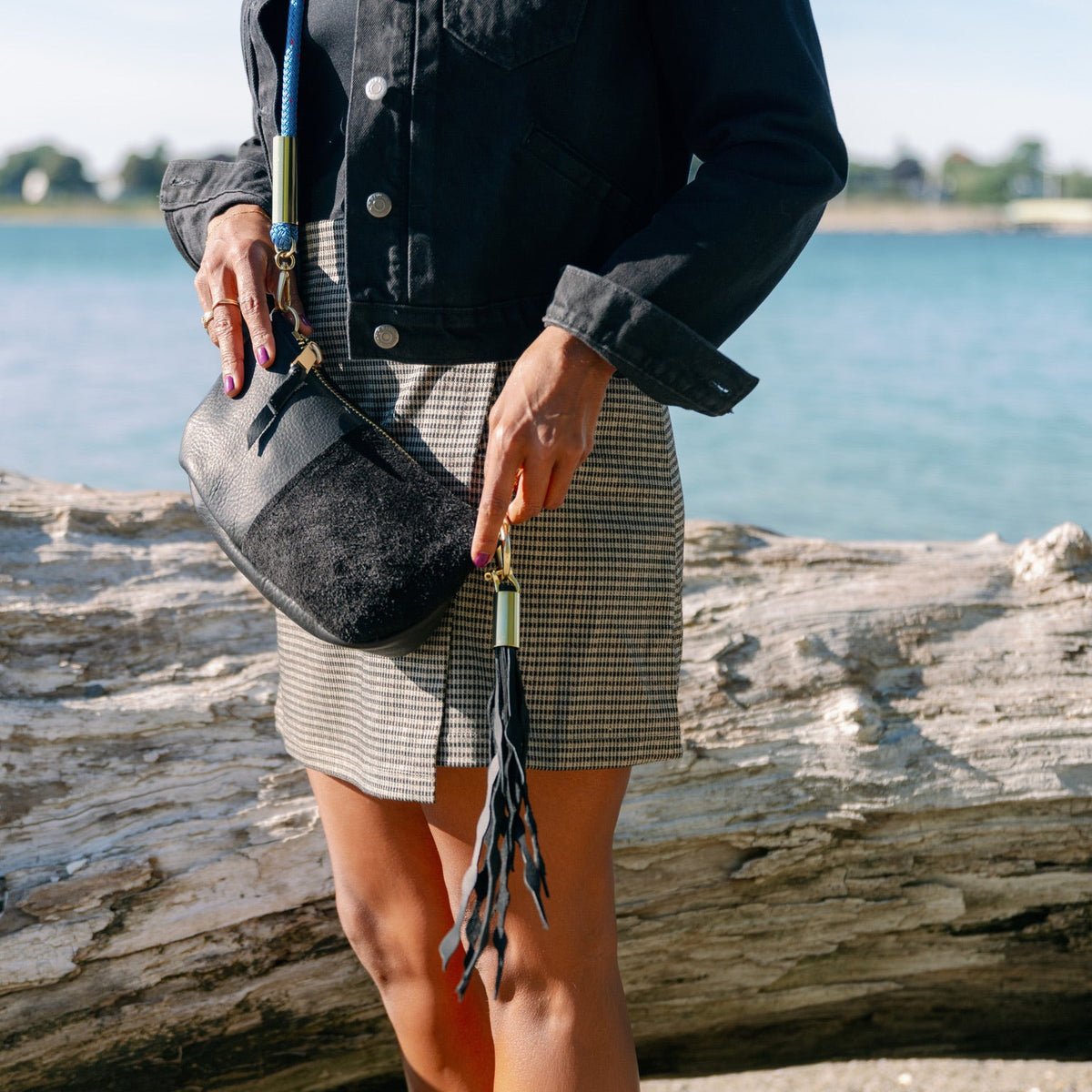 A person standing outdoors by the water holds a black crossbody bag with a tassel charm. The person is wearing a dark denim jacket and a gray-and-white checkered mini skirt.