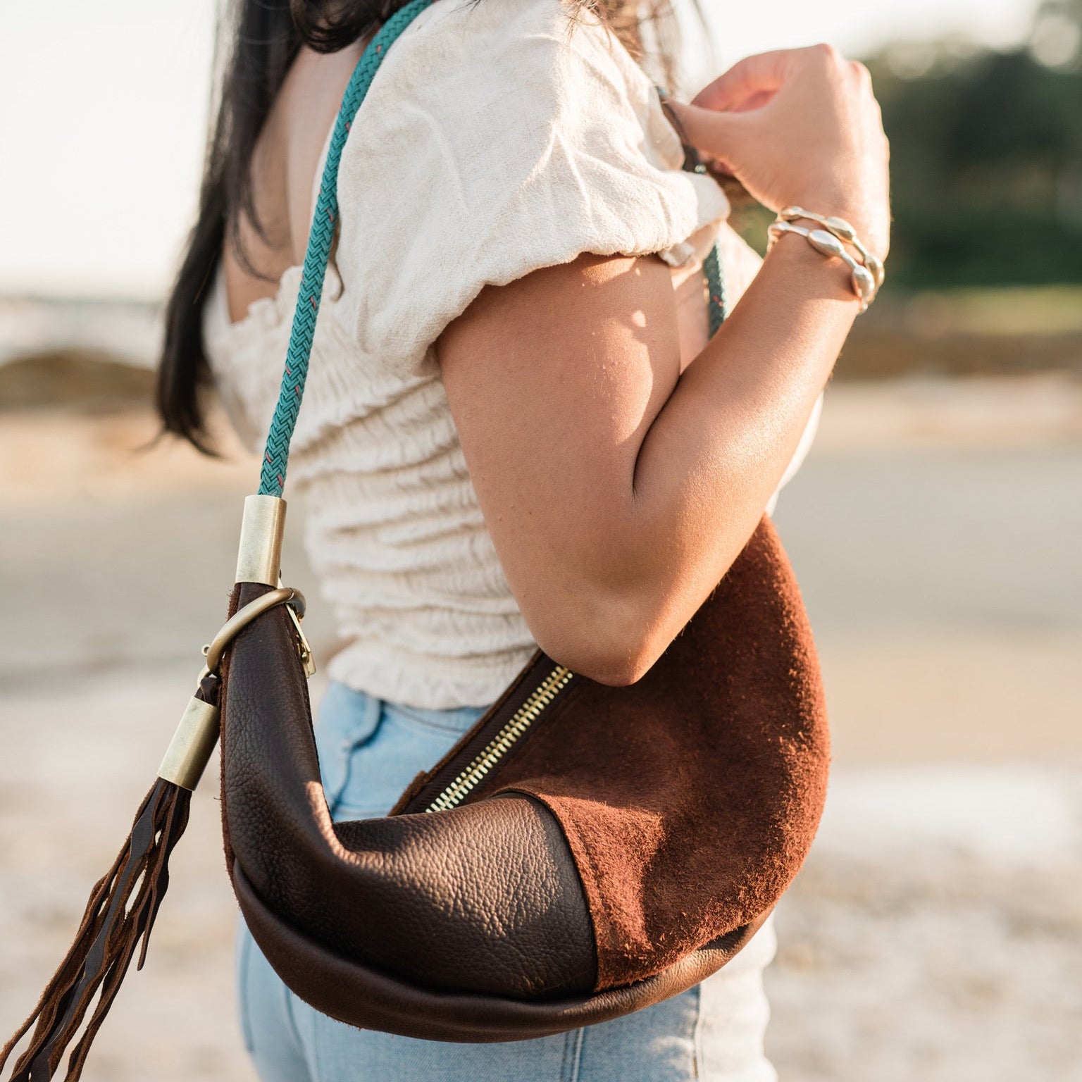 Woman holding a brown leather handbag with a strap over her shoulder on a beach.
