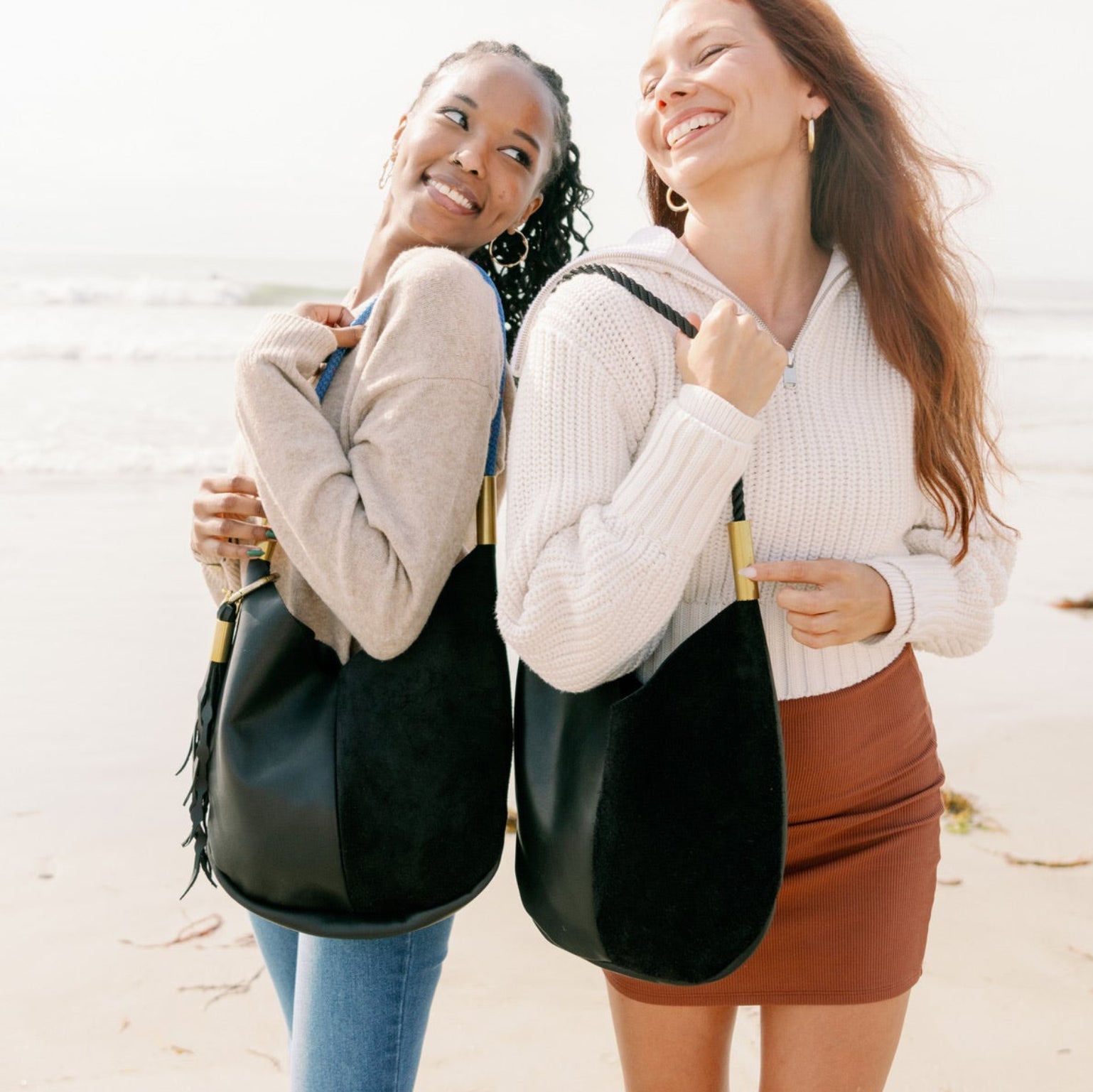Two women standing on a beach with black bags over their shoulders.