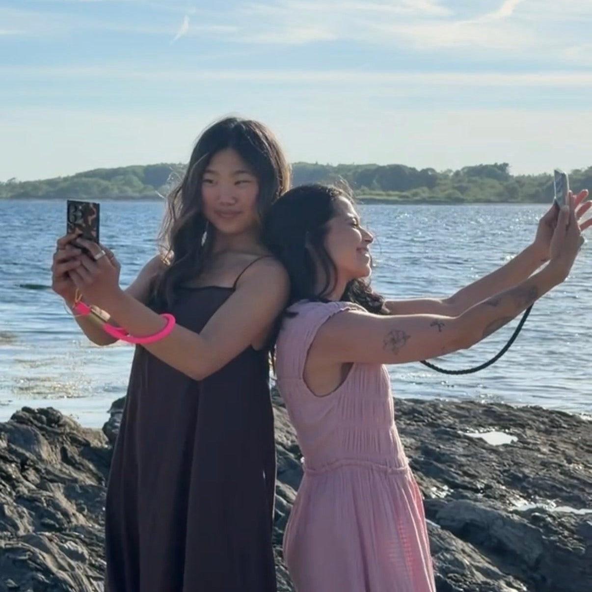 Two women taking a selfie on a rocky beach with a body of water and sky in the background.