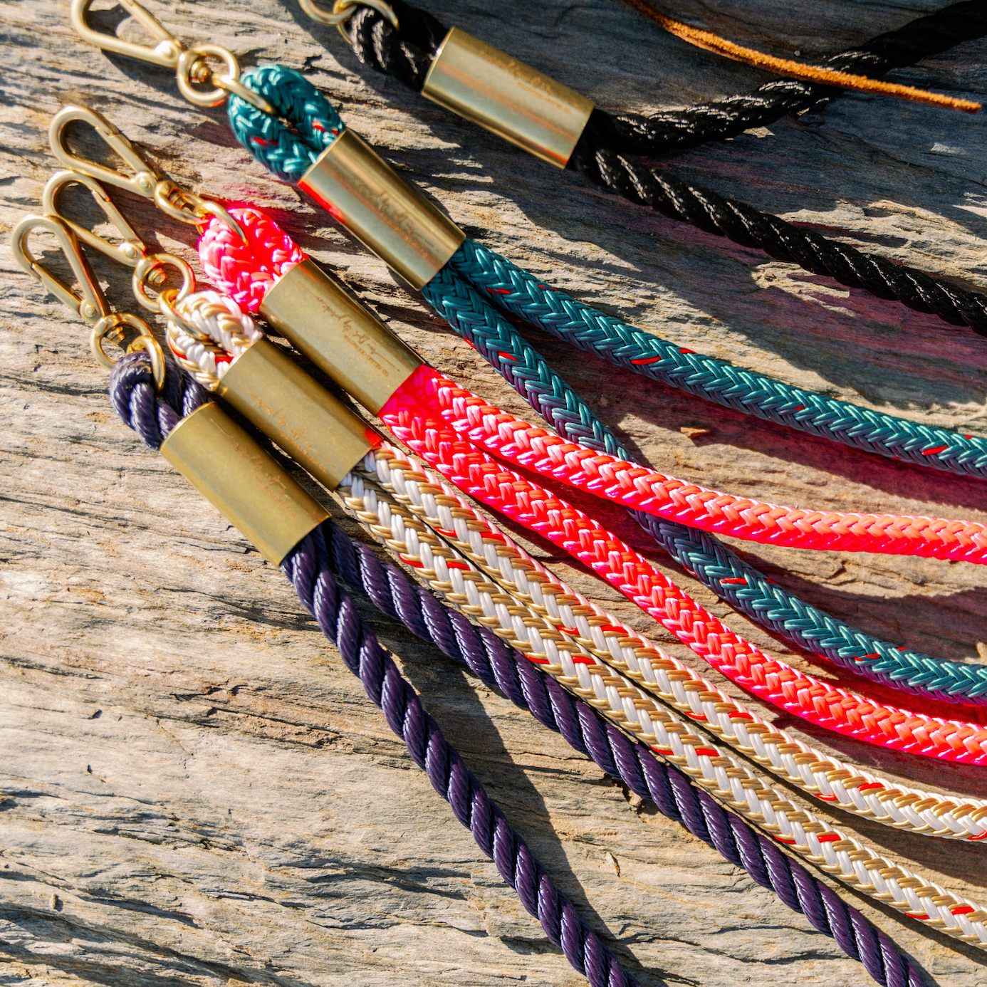 Five braided rope loops with brass hardware and swivel clips, laid out on a weathered stone surface. The loop wristlets include navy, cream, neon pink, teal and black ropes.