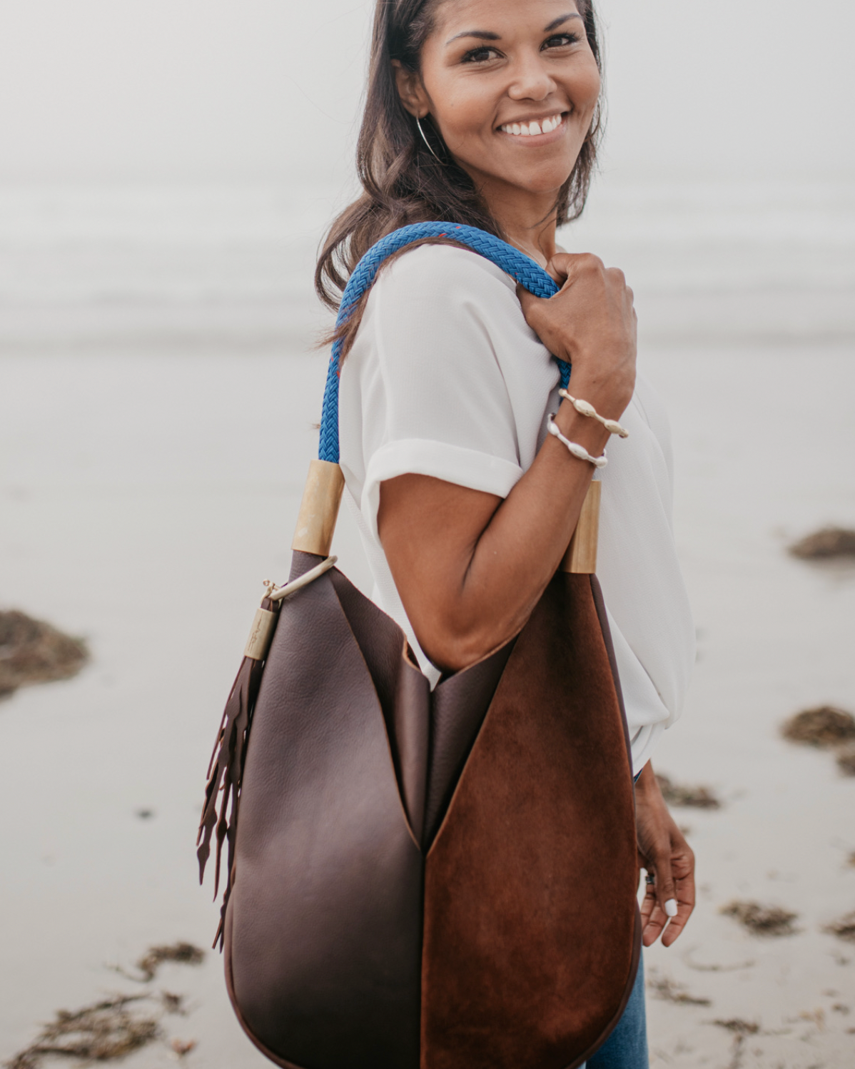 A woman smiling on a beach while carrying a large brown leather shoulder bag with a bright blue strap.