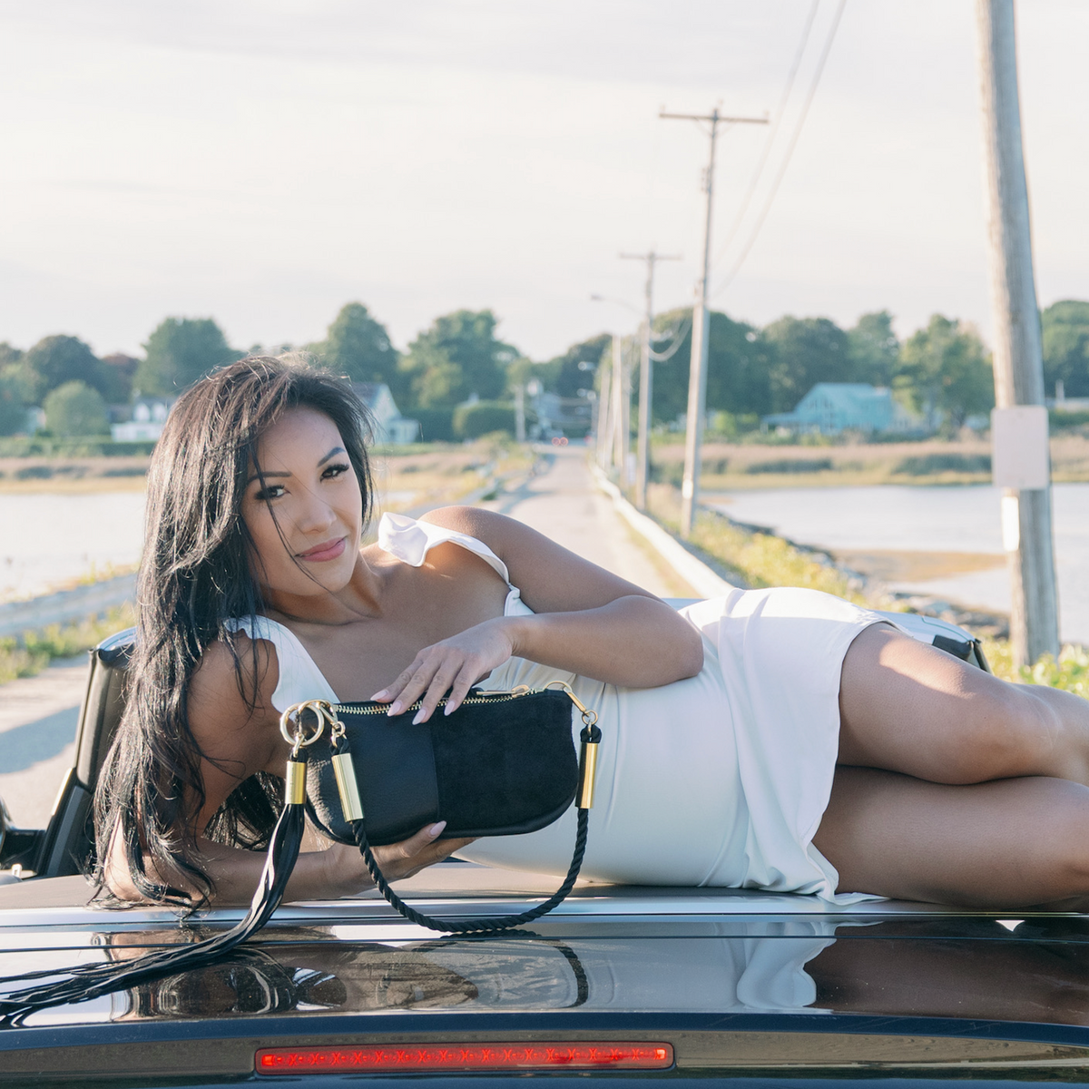 A woman in a white dress laying on the trunk of a black Audi car, holding a Black Classic Zip, with a lakeside road and trees in the background.