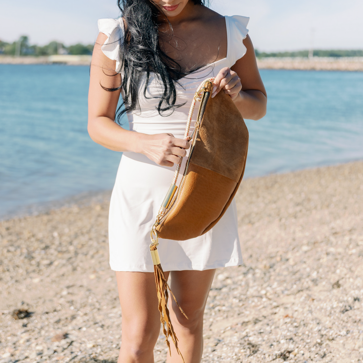 A woman wearing a white dress stands on a pebble beach zipping open the Beach Nut Sling with a matching Seaweed Tassel, with the blue ocean in the background.