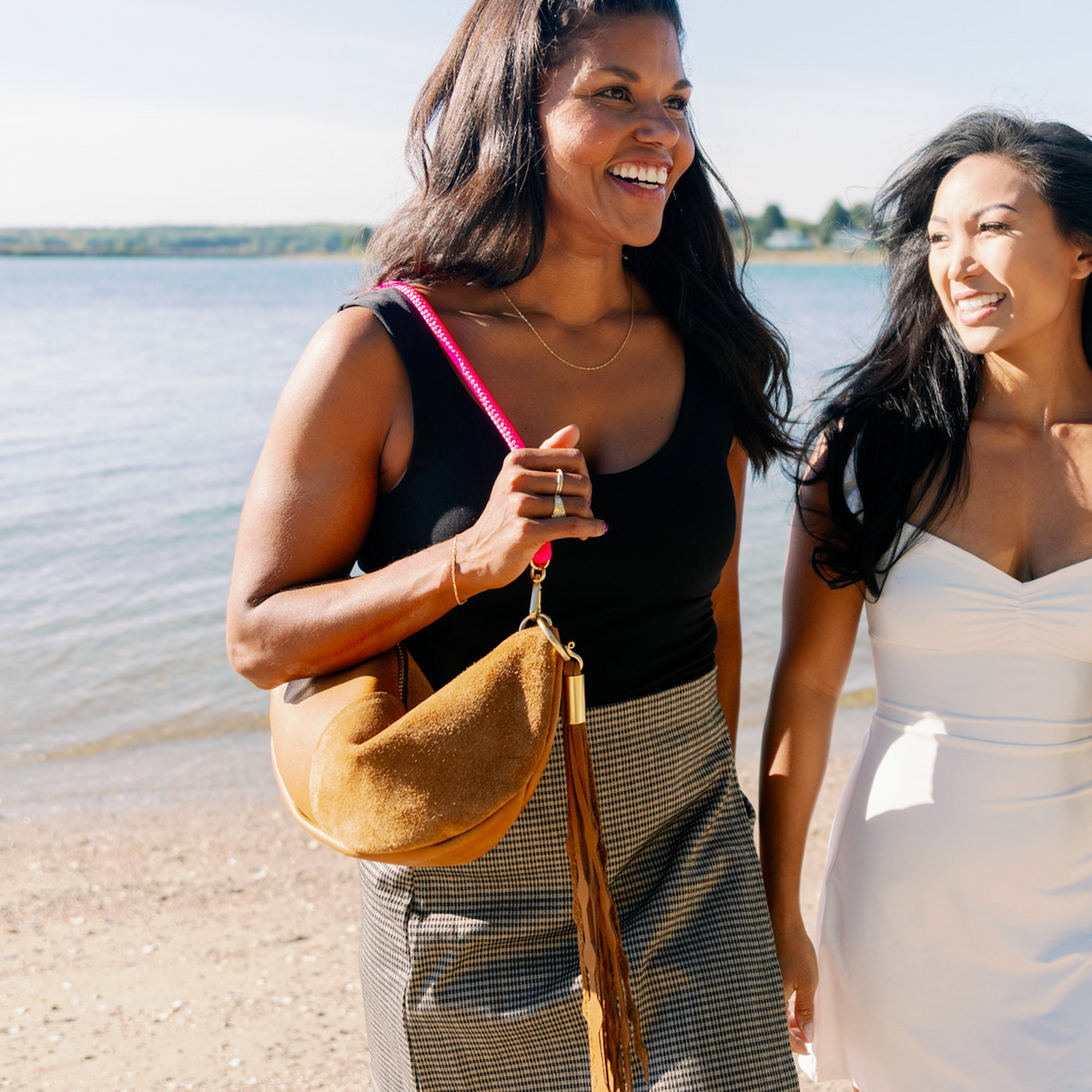Two women standing on a sandy beach next to the water, smiling and laughing. The woman on the left wears a black top, gray plaid skirt, and carries a Beach Nut Sling with a Neon Pink rope and a matching Dramatic Seaweed Tassel.