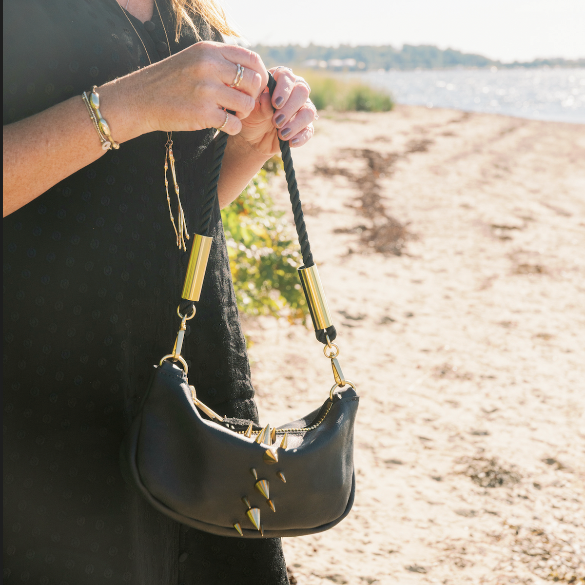 Becky at the beach holding a Black Convertible Sea Urchin Bag with the Summer Night Black rope, she is wearing a black dress and gold Seaweed Jewelry.