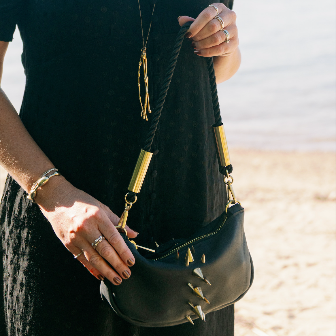 A woman in a black dress holding a Black Sea Urchin Bag with brass spikes, photographed outdoors near water and sand.