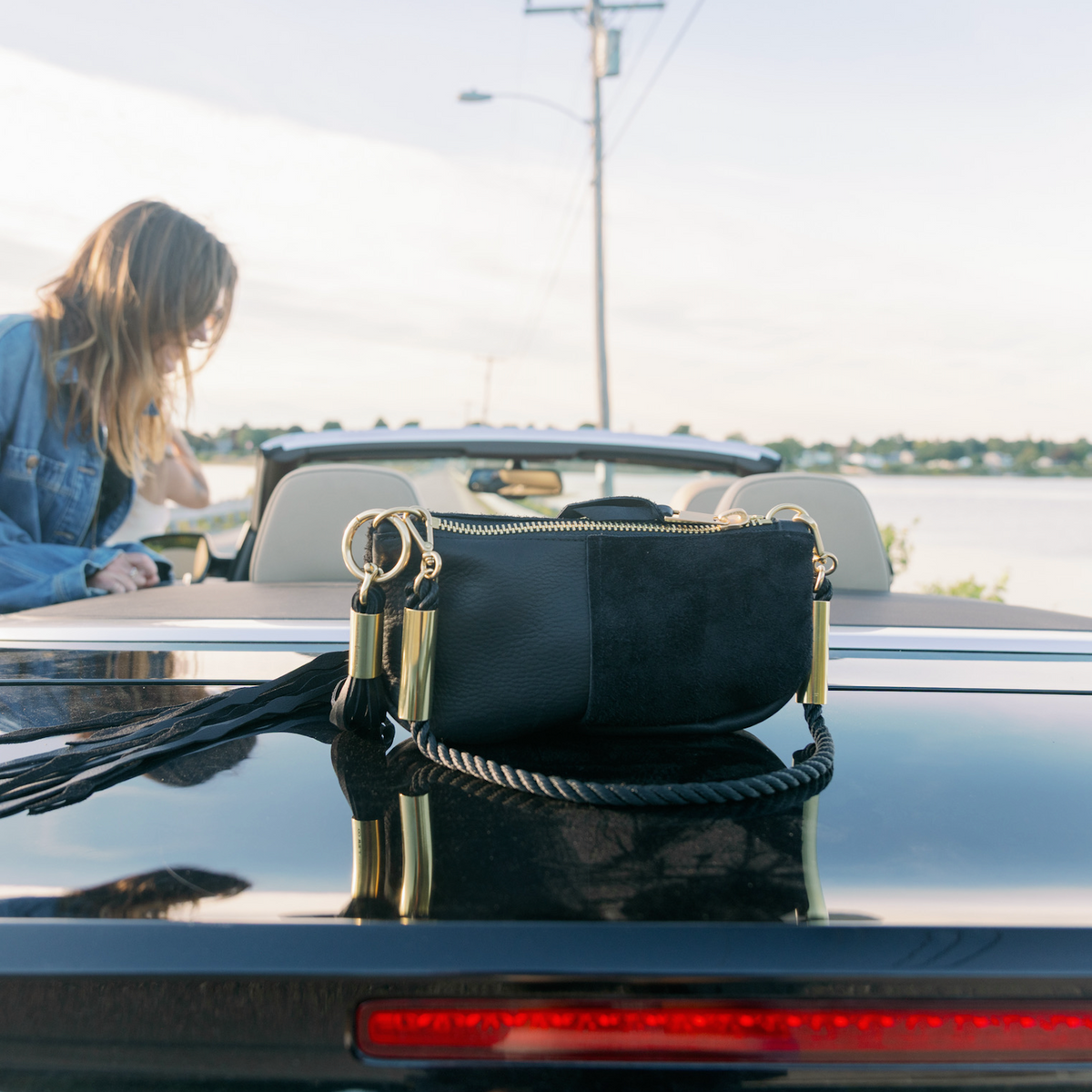 A Black Classic Zip bag with a Summer Night Black rope sitting on the trunk of a black convertible car, with a woman in a denim jacket leaning on a convertible nearby by a waterfront.