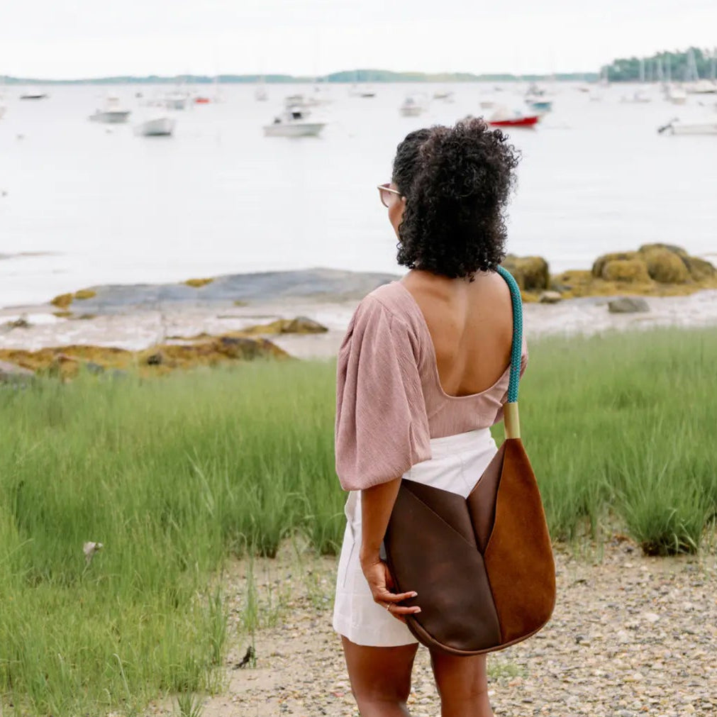 Woman standing on a path by a body of water with boats, wearing a brown bag.