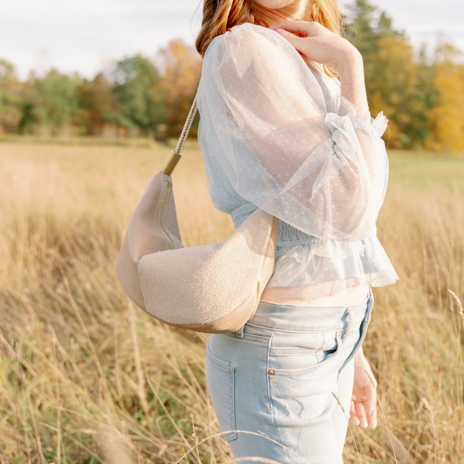 Woman holding a beige handbag in a field with trees in the background