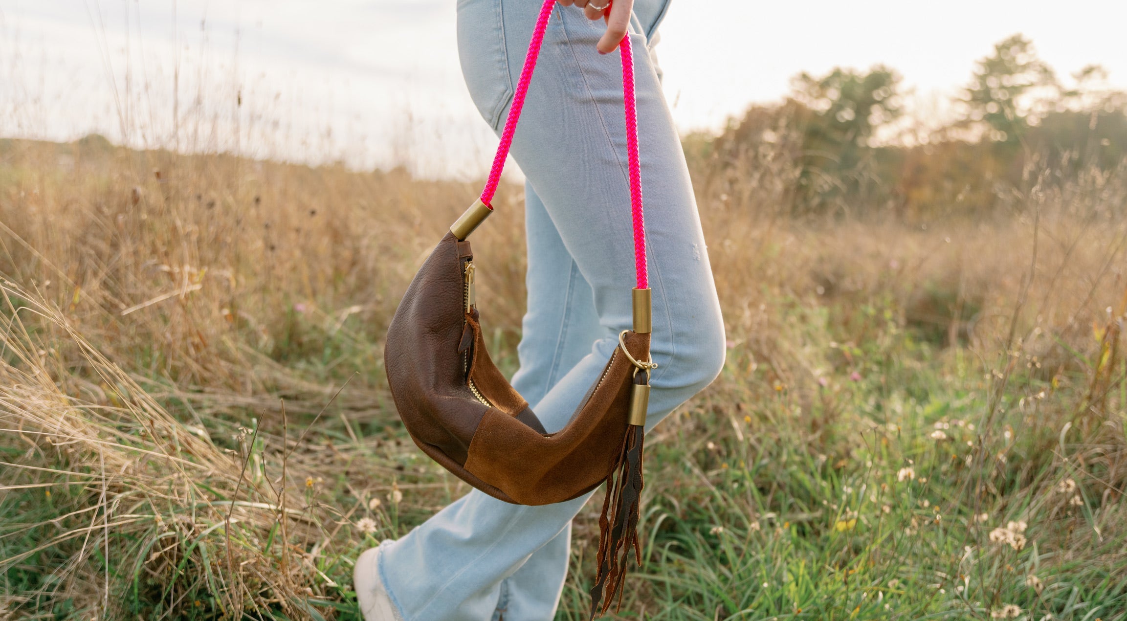 Person holding a brown leather handbag with a pink strap in a field