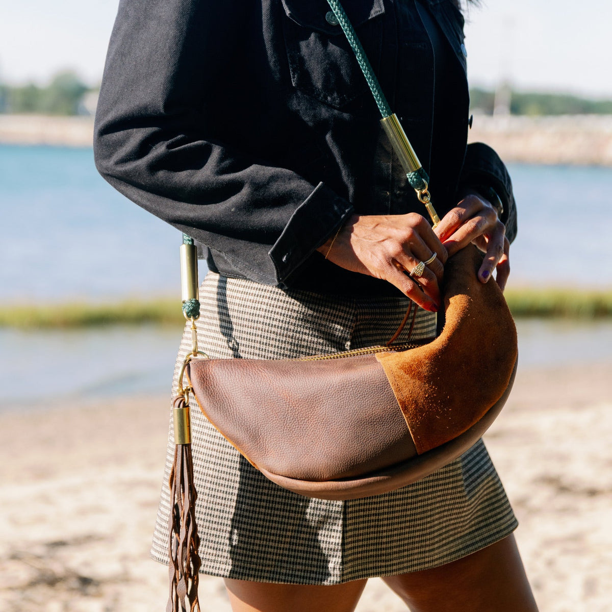 A person standing on a sandy beach wearing the Brown Sling Bag with a matching Seaweed Tassel and the Seaside Teal rope, paired with a black jacket and a gray-and-white patterned skirt.