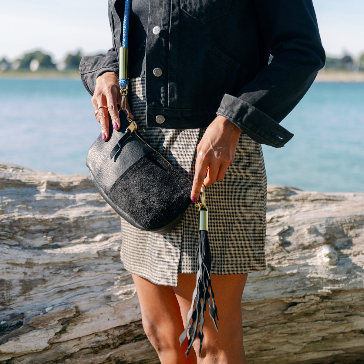 A person standing outdoors by the water wears a black denim jacket, a gray and white houndstooth mini skirt, and carries a Black Classic Zip bag with a Seaweed Tassel.