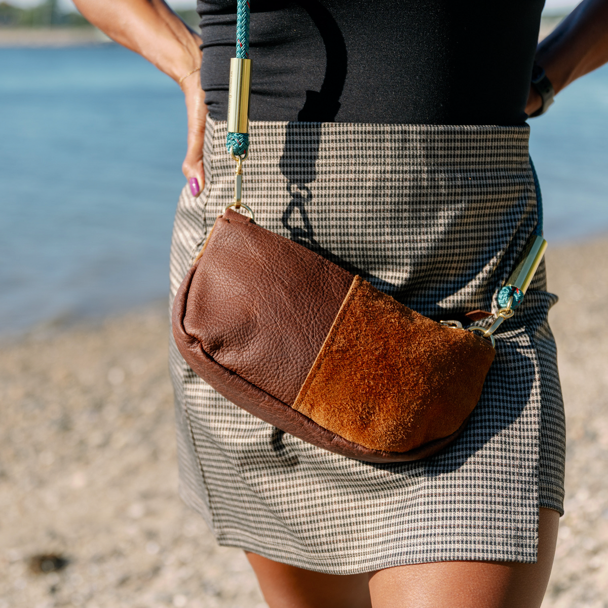 A Brown Classic Zip bag with a Seaside Teal rope, worn by a model standing on a pebble beach with water in the background.