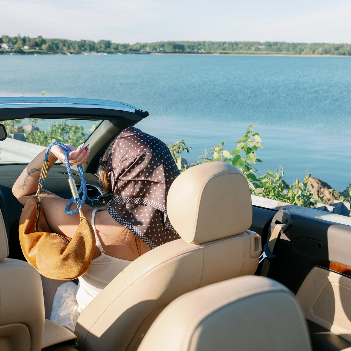 A woman sits inside a beige interior convertible car with the top down, parked beside a large blue lake. She is holding the Beach Nut Sling Bag with the Harborside Blue rope.